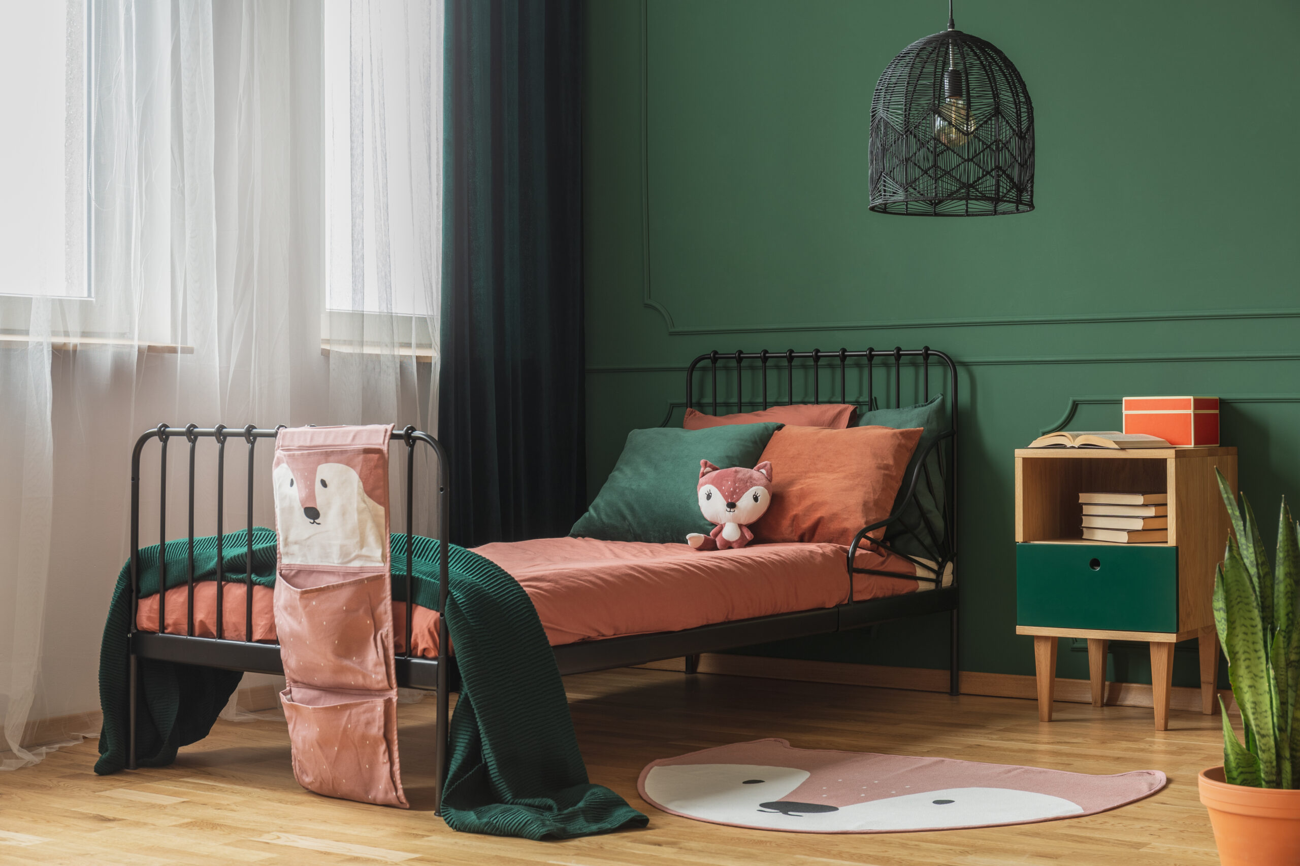 Real photo of a rug shaped like a fox on the wooden floor of a child's bedroom interior with orange sheets and pillows on a black bed standing next to the window
